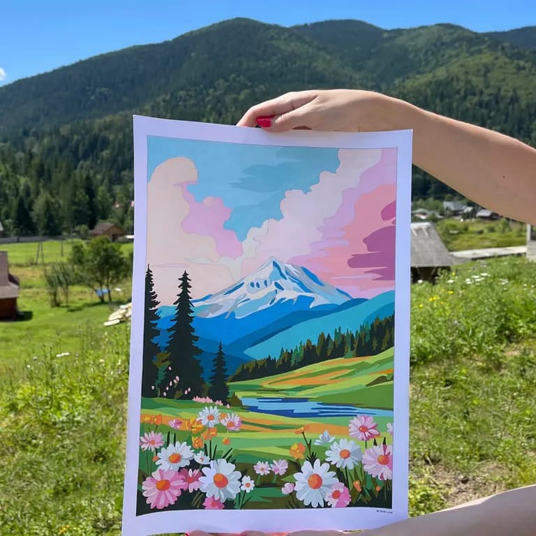 Hands holding a finished mountain landscape coloring page against a real mountain valley backdrop